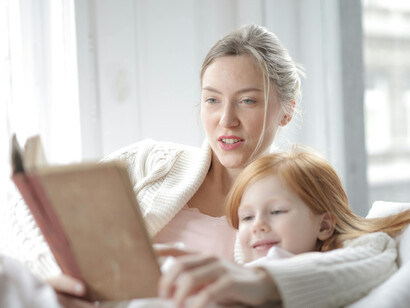 A mother and daughter lie in bed, reading a book together, fully engaged and sharing a warm, intimate moment
