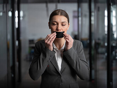 A woman in a gray suit stands in a sleek office, her mouth taped shut in silence, expressing concern over political persecution in an authoritarian regime