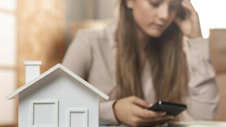 A woman holds her phone while standing behind a miniature wooden house, deep in thought