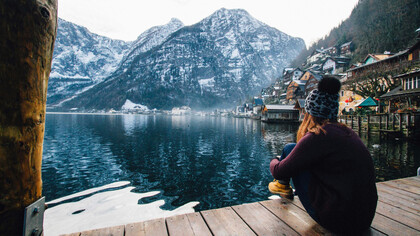 A woman sits on a wooden pier, gazing out at a serene winter landscape of a lake and snow-capped mountains