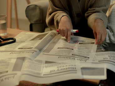 A graduate sits at home, thoughtfully scanning job listings in a newspaper