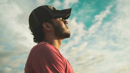 A man in a black cap stands with his eyes closed beneath a cloudy sky, lost in thought