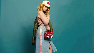 Foto de mulher vestindo saia jeans, agasalho de lã com uma jaqueta verde musgo e um boné branco