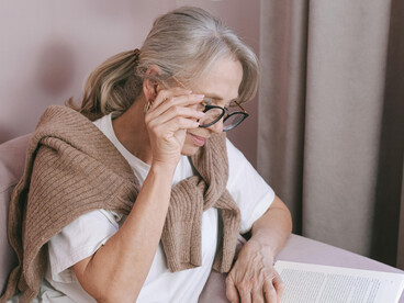 Elderly woman with glasses relaxing on a couch while reading a book at home