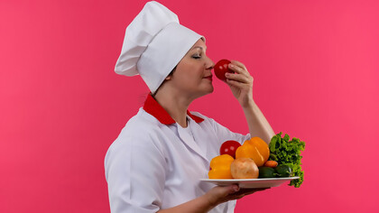A middle-aged female chef in a chef's uniform, holding a plate of vegetables, sniffing a tomato in her hand