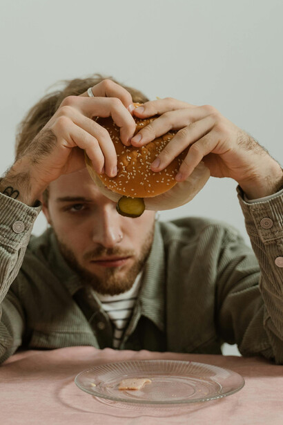 A man sits with his hand on his head, staring at a burger on a table in a minimalist studio, appearing depressed