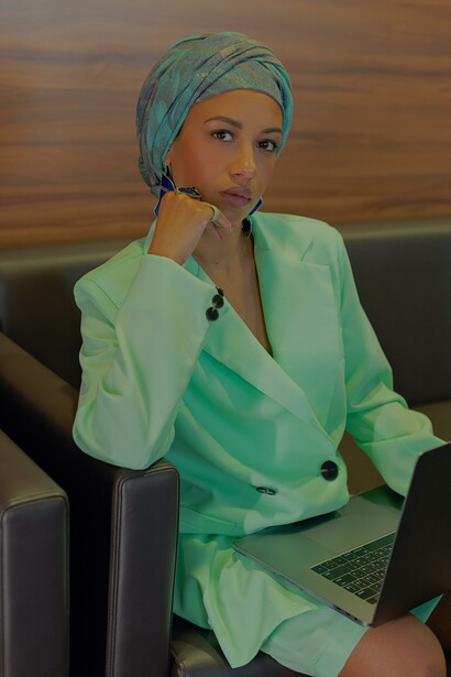 Uma mulher empreendedora de blazer verde sentada no sofá com a mão no queixo