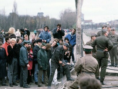 A crowd gathers on the West German side of the Berlin Wall at Potsdamer Platz as the structure is dismantled, while an East German policeman gestures toward the onlooker