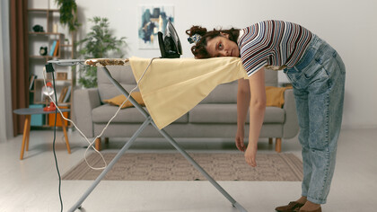 A woman leans on an ironing board, visibly exhausted from housework