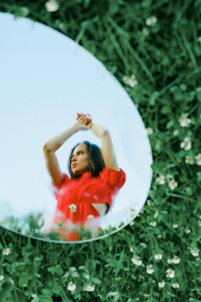 The mirror’s reflection captures her beautiful face, a visual metaphor for the tension between modern self-awareness and the timeless quest for authenticity