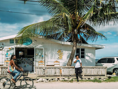 In the vibrant sunlight of Negril, Jamaica, a man sporting a sleek black shirt gracefully pedals his bicycle, creating a harmonious image against the backdrop of a swaying palm tree