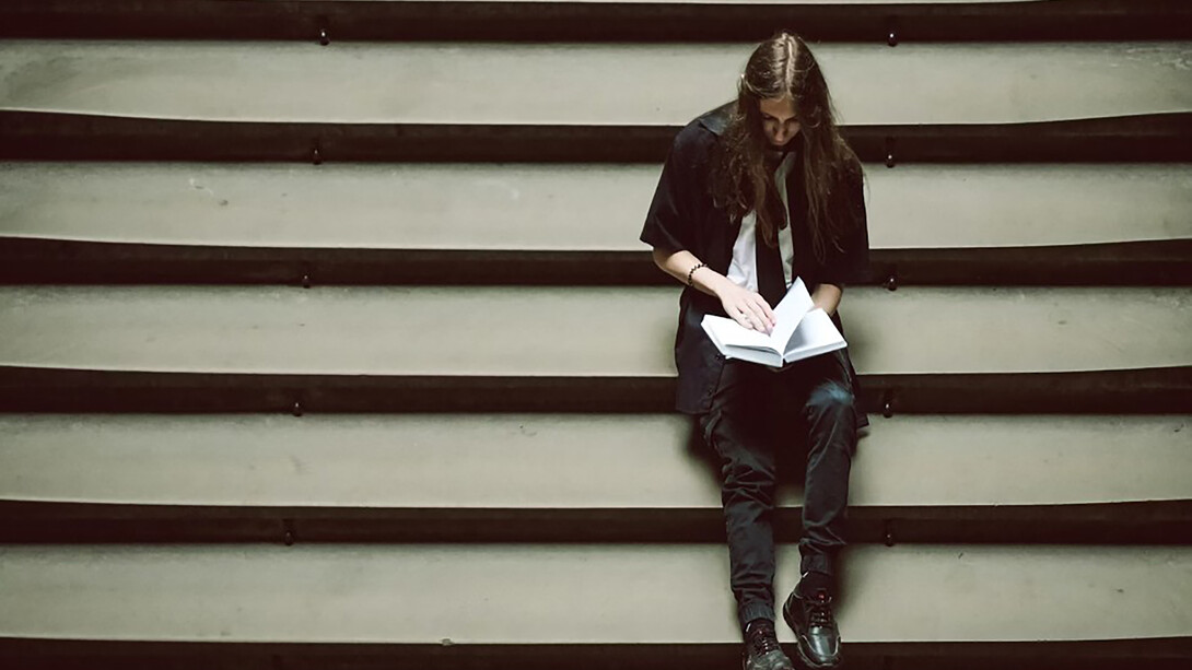 A person sitting on a set of stairs, holding a book, lost in thought or overwhelmed by the weight of their studies