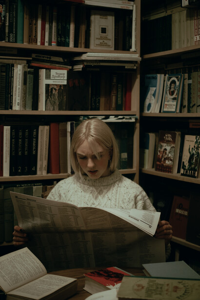 A young woman, surprised, reading a newspaper, in a library, with books all around her