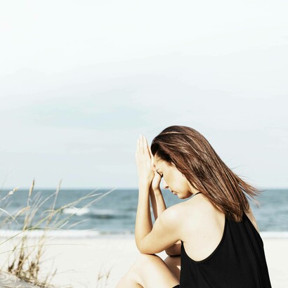 Donna seduta in spiaggia, con le mani giunte davanti alla fronte in un atto di preghiera