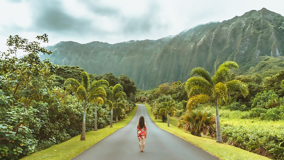 A woman enjoys a leisurely stroll along the black concrete road amidst the scenic backdrop of Ho’omaluhia Botanical Gardens in Oahu, offering serene views of majestic mountains