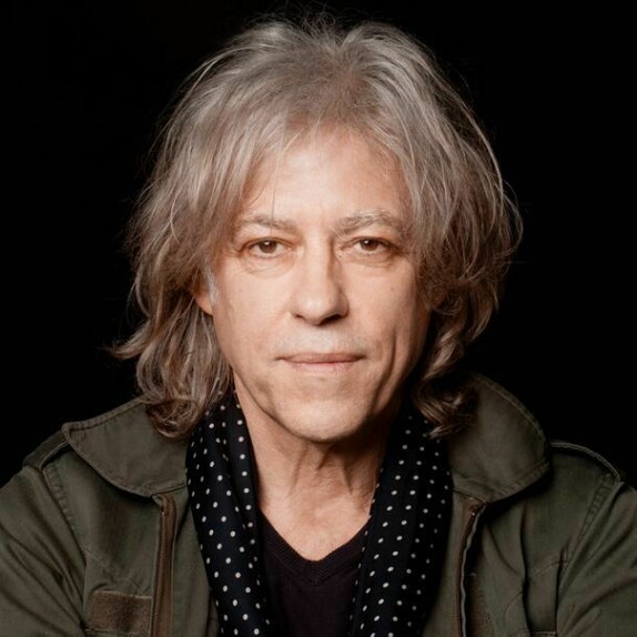 Bob Geldof, grey hair to his shoulders, in a green jacket and spotted scarf against black