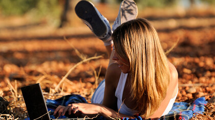 A woman working on her laptop while lying on a bed of yellow autumn leaves, balancing self-care with work