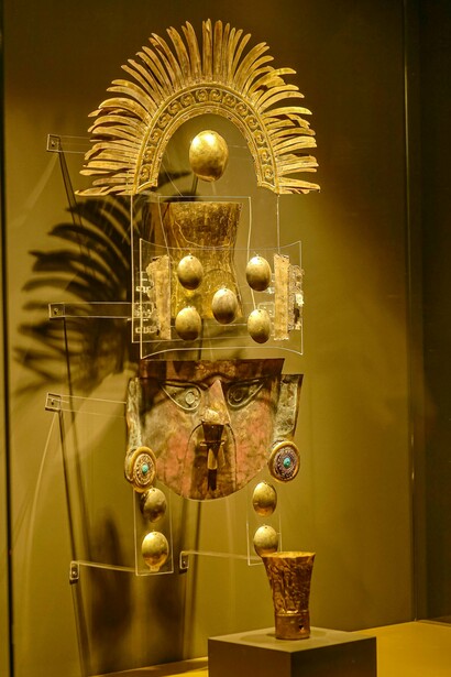 Adornos cerimoniais de culturas antigas exibidos em vitrines iluminadas. Para além da preservação material, cresce o questionamento sobre quem deve guardar objetos carregados de memória e identidade