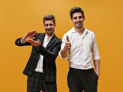 Two confident young businessmen with dark hair, dressed in classic white shirts, celebrate excitedly against a vibrant orange background