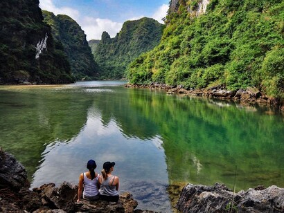 Two women are sitting by the lake in Ninh Bình, Vietnam, enjoying the serene view of the water surrounded by lush green mountains and misty hills
