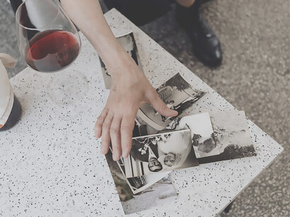 A woman, cradling a wine glass in her hand, gazes wistfully at photographs, her eyes reflecting a poignant mix of sadness and longing