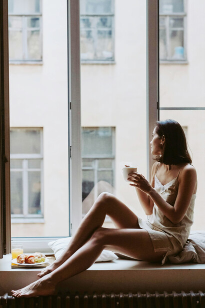 A contemplative woman enjoys a book and coffee by the window, gazing into the distance as sunlight streams through the glass