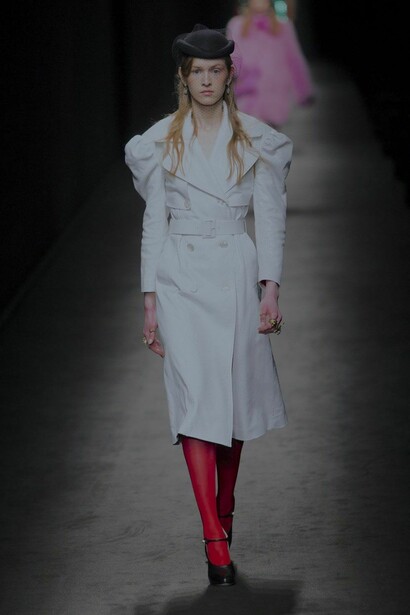 Modelo veste meia calça vermelha com salto, sobretudo branco e chapéu preto