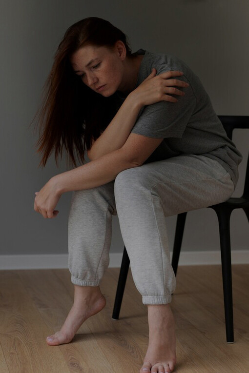 A woman sits alone on a chair with a cloud over her head, her expression reflecting deep anxiety and sadness
