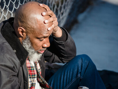 A middle aged black black sitting with his back on a fence, holding his head, eyes closed