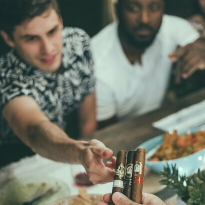 A group of friends gathers around a table, engaged in conversation as a hand extends three fine cigars, capturing the essence of cigar culture—a shared experience that transcends backgrounds and social status, bringing together those who appreciate the ritual and flavor of a good smoke