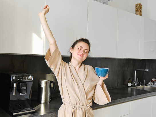 Brunette woman dancing and stretching in the kitchen while drinking coffee