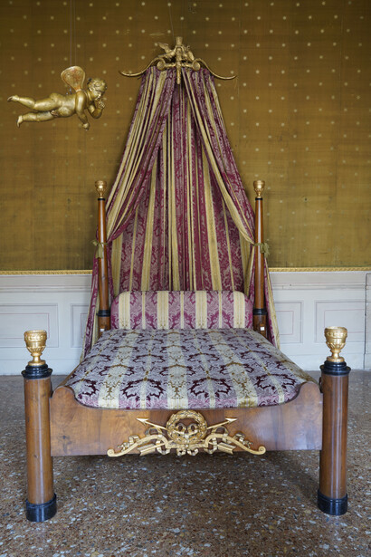 Letto stile Impero, primi decenni sec. XIX, di manifattura emiliana; sui montanti della testata due colonne lisce, sui montanti della pediera e sulle colonne lisce quattro vasi intagliati in legno dorati. Sul fronte della pediera è applicato intaglio dorato in legno con arco, frecce, corona d’alloro e due colombe. Baldacchino intagliato con arco, frecce e colombe, con stoffa rigata rossa e oro.