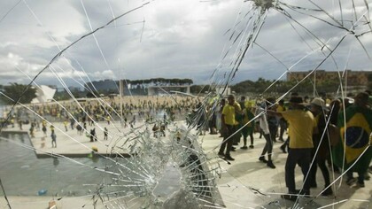 Por trás do vidro estilhaçado, manifestantes vestidos predominantemente de verde e amarelo ocupam a rampa de acesso ao Palácio do Planalto, durante os atos de 8 de janeiro de 2023. A destruição das sedes dos Três Poderes expôs a gravidade da ofensiva contra as instituições democráticas brasileiras perpetradas por estes mesmos representantes da extrema-direita