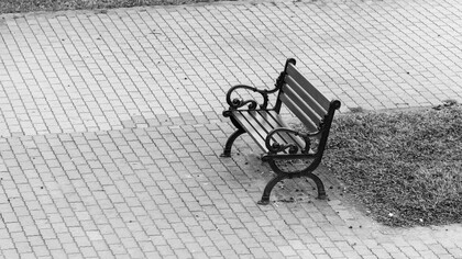 Um banco de parque solitário e vazio, capturado em preto e branco, permanece isolado em uma praça cercado pelo silêncio dos paralelepípedos em um ambiente urbano à espera de alguém que já não chega. Há ausências que ocupam mais espaço do que qualquer presença. Entre pedras e silêncio, um banco desocupado guarda memórias invisíveis. Algumas histórias continuam sentadas mesmo depois de quem as viveu partir