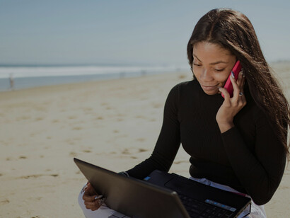 By the shoreline, a woman in a black long-sleeve shirt types on her laptop, capturing the essence of remote work and freedom