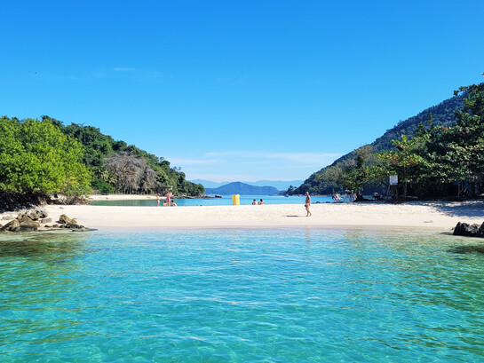 A praia aparece entre o mar, a vegetação e as montanhas ao fundo, formando uma paisagem de águas tranquilas e natureza exuberante. Em Ilha Grande, o afastamento do ritmo urbano abre espaço para desacelerar e reencontrar outros modos de estar no mundo