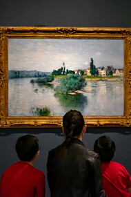 A woman in a black jacket sits on a red sofa with two children inside an art museum
