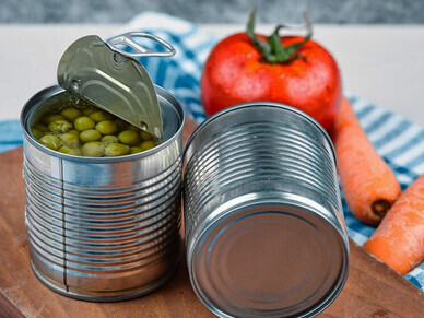 On a tablecloth, two cans of boiled green peas and a variety of fresh vegetables are displayed