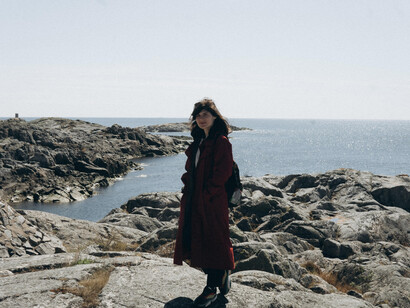 A solitary woman on a rugged shoreline, her long coat moving with the wind as she stands in the quiet tension of a transition phase