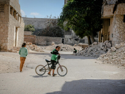 A child stands beside a bicycle amid the ruins of a demolished city in Idlib, Idlib Governorate, Syria