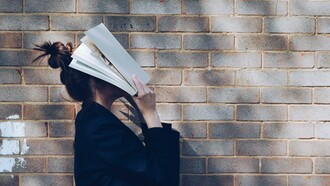 A schoolgirl stands against a rustic red brick wall, holding a book up to cover her face, reflecting student burnout