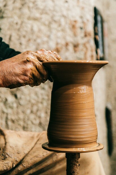 Man working with clay to form a vase in a naturally lit artist studio with craft supplies on the desk