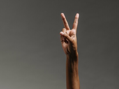 Uma mão reproduz o conhecido sinal de paz ou vitória com dois dedos levantados. Gestos como esse integram um repertório de sinais compartilhados socialmente, cujo significado é construído para além da linguagem verbal