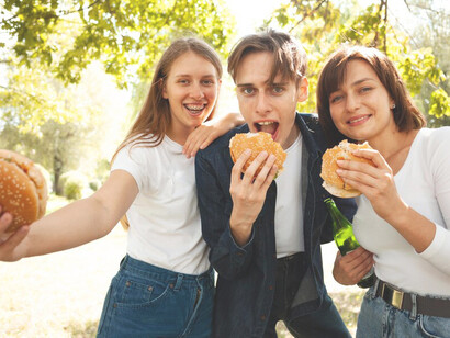 Friends posing with burgers and beer in the park, capturing the moment with selfies