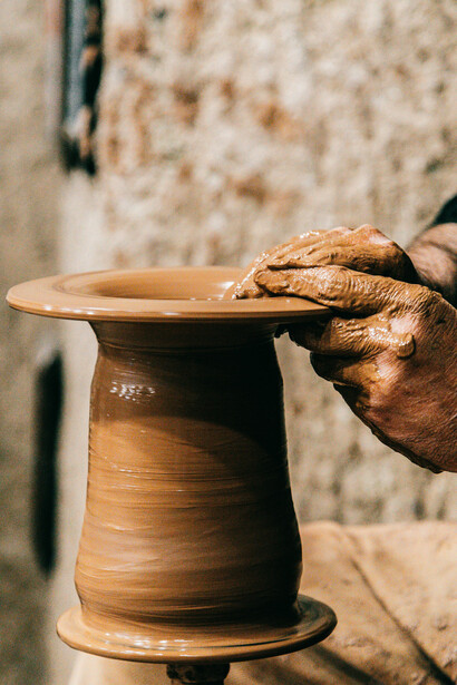 A man shapes a clay vase in his sunlit studio, with craft tools and materials spread across the desk