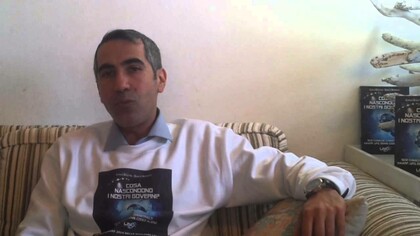 Carlo Di Litta resting comfortably on his sofa wearing a sweater that has an image of the cover of his book  "Cosa Nascondono I Nostri Governi?"