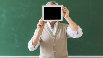 Un professore tiene un tablet con lo schermo nero nascondendo il viso, sullo sfondo una grande lavagna verde 