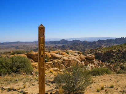 The vast and rocky terrain of Pioneertown Mountains Preserve is showcased beyond a weathered trail marker under a clear sky