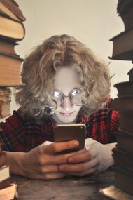 A young boy scrolling, glued to his smartphone while ignoring the piles of old books in front of him; an image that reflects the reality of the youth in our current generation, all about their smartphones and movie platforms, rejecting the good old novels