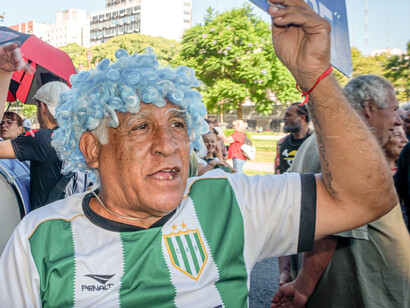 Manifestante luciendo una peluca rizada de color azul celeste y una camiseta deportiva verde y blanca mientras sostiene un cartel en una multitud de calle